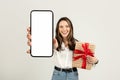 Joyful woman presenting a smartphone with a blank screen in one hand and holding a gift Royalty Free Stock Photo