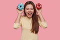 Joyful excited lady exclaims from happiness, carries two blue and red donuts, has tempatation to eat it, keeps to diet