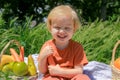 joyful fair-haired child holds a carrot and smiles, basket with fruits on green grass