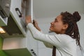 Joyful female with braids opening cupboard indoor, woman opens kitchen cabinet at home Royalty Free Stock Photo