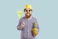 Joyful funny bearded chubby man with pineapple and insect net on light blue summer background.