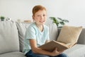 Joyful ginger boy sitting on couch at home, reading book Royalty Free Stock Photo