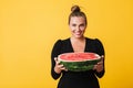 Joyful girl in black dress happily looking in camera holding half of big watermelon in hands over yellow background