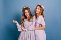 Joyful girl in circlet of flowers smiling to camera during photoshoot with friend. Studio portrait of adorable blonde Royalty Free Stock Photo