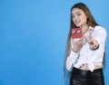 Joyful girl in white shirt demonstrating teeny red gift box, while showing victory gesture indoors.