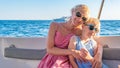 Joyful girls sit on a yacht, young with delicate lips, adult hugs her daughter. Around the sea, enjoy in sunglasses in