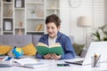 Joyful guy schoolboy reads a book in his room at the table, does homework, works with a tutor online on a laptop Royalty Free Stock Photo