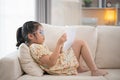 Joyful happy asian child baby girl smiling and reading book while sitting on couch sofa in living room at home. Girl relex reading Royalty Free Stock Photo