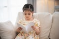 Joyful happy asian child baby girl smiling and reading book while sitting on couch sofa in living room at home. Girl relex reading Royalty Free Stock Photo