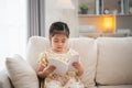 Joyful happy asian child baby girl smiling and reading book while sitting on couch sofa in living room at home. Girl relex reading Royalty Free Stock Photo