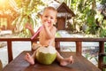 A joyful and happy little one year old child sits on a wooden table and drinks coconut milk from fresh green coconut through a Royalty Free Stock Photo