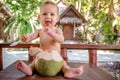 A joyful and happy little one year old child sits on a wooden table and drinks coconut milk from fresh green coconut through a Royalty Free Stock Photo