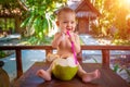 A joyful and happy little one year old child sits on a wooden table and drinks coconut milk from fresh green coconut through a Royalty Free Stock Photo