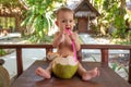 A joyful and happy little one year old child sits on a wooden table and drinks coconut milk from fresh green coconut through a Royalty Free Stock Photo