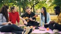 Joyful hipster is singing and playing the guitar sitting on blanket in park with friends and having fun, people are Royalty Free Stock Photo