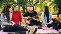 Joyful hipster is singing and playing the guitar sitting on blanket in park with friends and having fun, people are