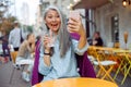 Joyful hoary haired Asian lady takes selfie with smartphone on outdoors cafe terrace