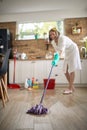 Joyful housewife cleaning the kitchen, wet mopping the floor, with chemical bottles in the background