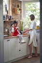 Joyful housewife woman washing dishes in the kitchen at daytime, with her daughter sitting on kitchen counter eating a muffin,