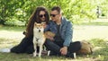 Husband and wife are using smartphone resting on lawn in park, watching screen and talking and fussing dog. Modern