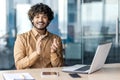 Joyful indian man in casual wear clapping hands while sitting at workplace with laptop in modern office. Young male with