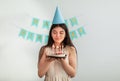 Joyful Indian teen girl in birthday hat blowing candles on cake at home