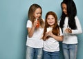 Joyful kids girls friends in blue jeans and white t-shirts stand with glasses of water, milk and juice in hands