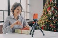 Joyful lady sitting at table with Xmas tree on background in front of phone on tripod