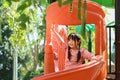 Joyful little asian girl having fun on playground in public park surrounded by green trees at sunlight morning