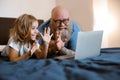 Joyful little girl with funny bearded daddy wave hands to laptop camera at videochat on bed Royalty Free Stock Photo