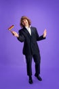 Joyful man in suit holding grogger symbol used to drown out name of Haman villainous character against purple backdrop.