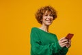Joyful middle-aged ginger woman using smartphone isolated over yellow background