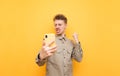 Joyful nerd with glasses, mustache and shirt stands on a yellow background with a smartphone, looks into the screen and rejoices Royalty Free Stock Photo