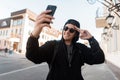 Joyful nice young hipster man in trendy hoodie hooded in dark sunglasses in a stylish cap with a smile makes the selfie outdoors