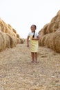 Joyful playful young girl with braid walking on hay country field road between stacks of hay looking at camera wearing Royalty Free Stock Photo