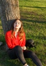 Joyful positive Asian girl enjoying weekend outdoors. Young woman sitting on red blanket, leaning back on tree, holding