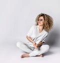 Cheerful positive blonde curly woman in white pants and blouse sits on the floor with crossed legs and dances