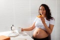 Joyful pregnant young beautiful woman eating croissant and drinking tea during morning breakfast. Concept of pleasant Royalty Free Stock Photo