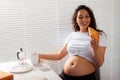 Joyful pregnant young beautiful woman eating croissant and drinking tea during morning breakfast. Concept of pleasant Royalty Free Stock Photo