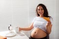 Joyful pregnant young beautiful woman eating croissant and drinking tea during morning breakfast. Concept of pleasant Royalty Free Stock Photo
