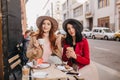 Joyful red-haired girl drinking tea while talking with friend. Brunette young woman spending time in restaurant with her Royalty Free Stock Photo