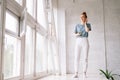 Joyful redhead young woman is using cell phone while standing near big panoramic window. Royalty Free Stock Photo