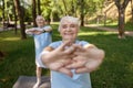 Joyful senior lady does sports exercise training with friend on lawn in city park