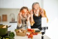 Joyful senior spouses filming content in kitchen for their food blog on smartphone on tripod, selective focus Royalty Free Stock Photo