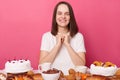 Joyful smiling delighted brown haired woman in white t shirt sitting at table with delicious desserts isolated over pink