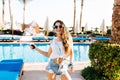 Joyful smiling girl in white shirt holding phone and dancing in front of swimming pool. Slim graceful young woman in Royalty Free Stock Photo