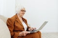 a joyful, sweet, gray-haired elderly woman in a brown suit is sitting on a beige sofa in a bright room working remotely