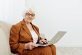 a joyful, sweet, gray-haired elderly woman in a brown suit is sitting on a beige sofa in a bright room working remotely