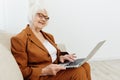 a joyful, sweet, gray-haired elderly woman in a brown suit is sitting on a beige sofa in a bright room working remotely