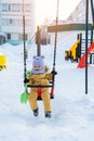 A joyful toddler in yellow warm clothes swings on a swing on a children`s playground in winter, front view